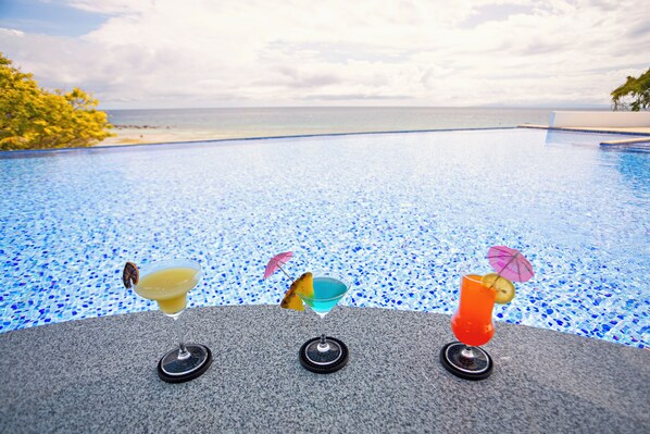 Vista a la piscina con tres cócteles sobre una superficie de granito, que da a una piscina azul cristalina y al océano.