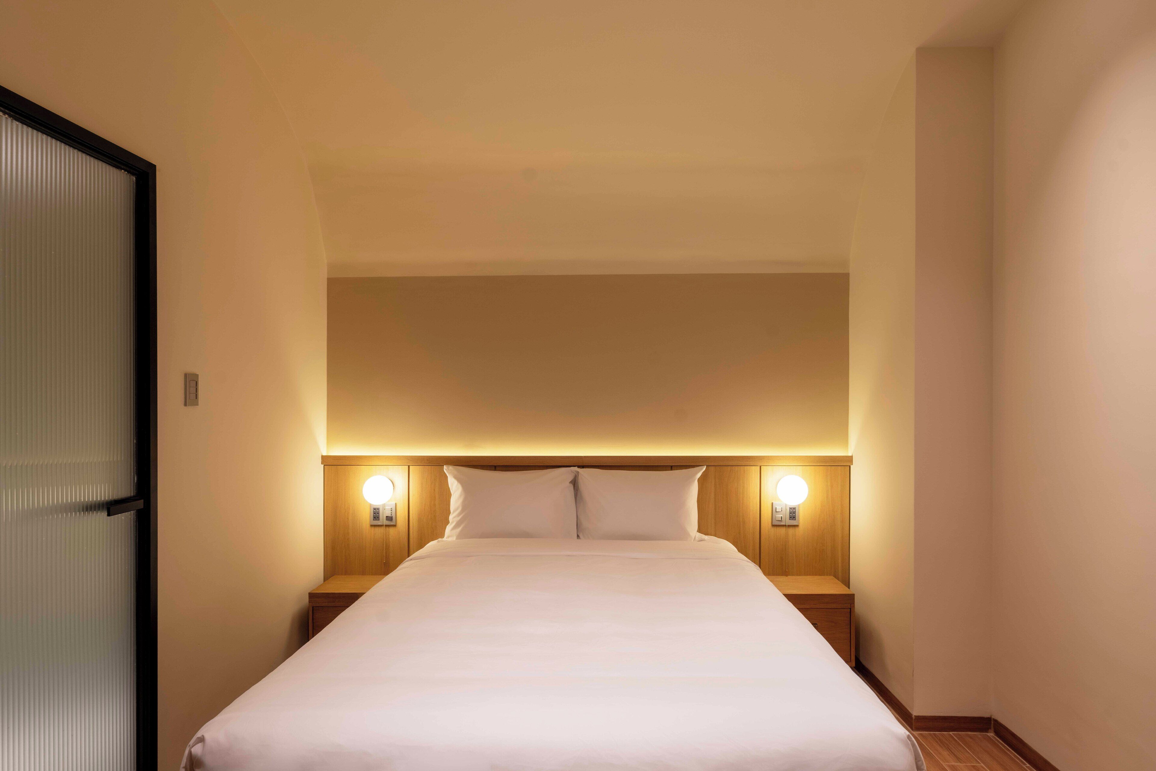Hotellihuone, jossa on suuri sänky, puinen pääty, yöpöytävalaisimet ja ovi, jossa on himmeälasista valmistettu paneeli.