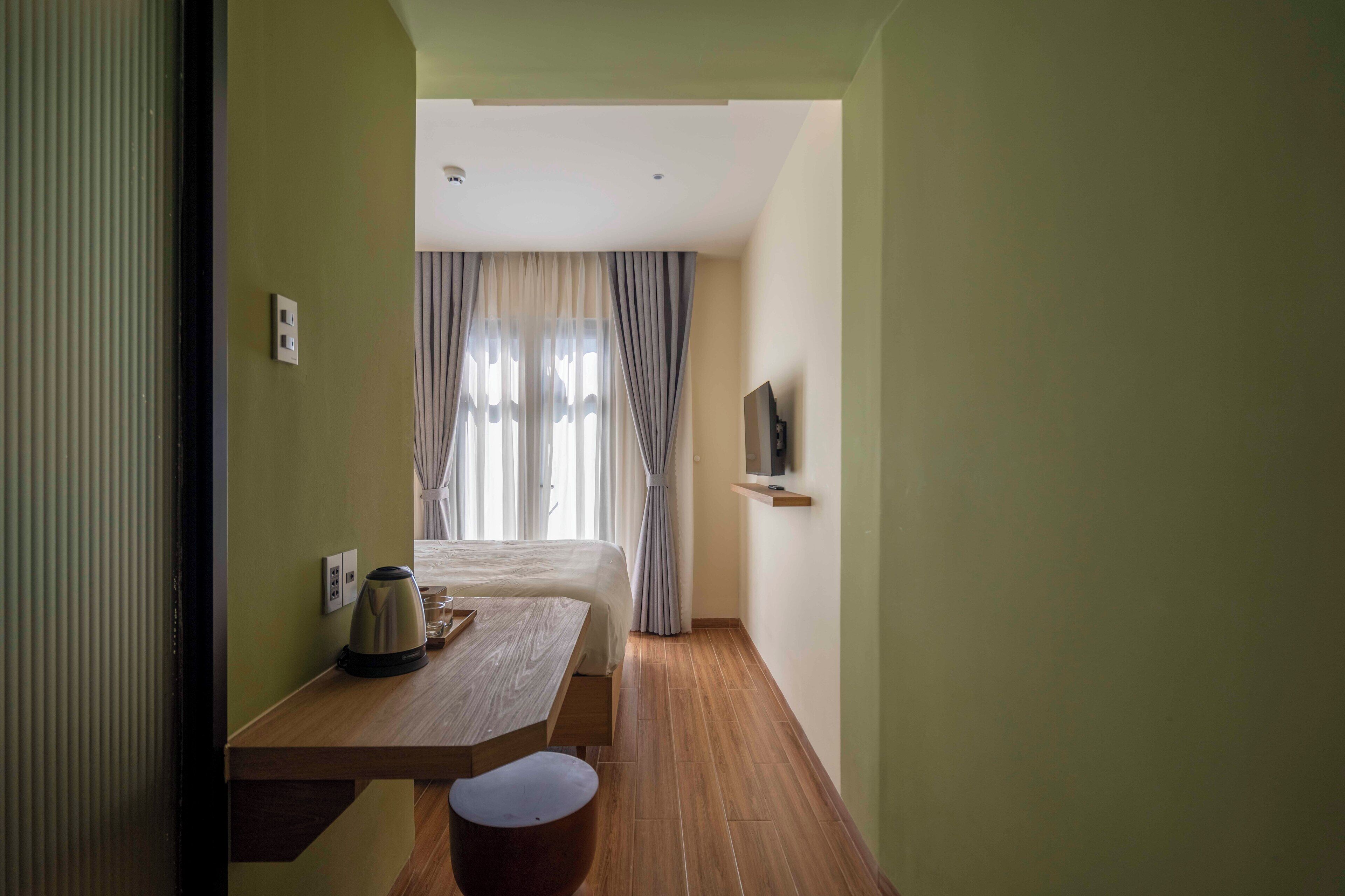 Une chambre d’hôtel avec un lit, une table avec une bouilloire, un téléviseur posé sur une étagère et des rideaux à la fenêtre.