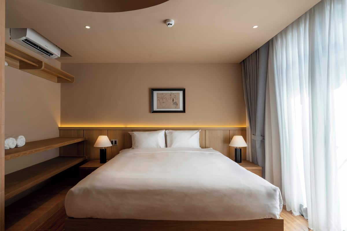 Une chambre d’hôtel moderne avec un grand lit, une tête de lit en bois, des lampes de chevet, une œuvre d’art encadrée au mur et une fenêtre avec des rideaux.