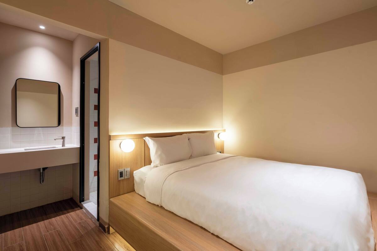 Une chambre d’hôtel avec une tête de lit en bois, un lit blanc, une petite table de chevet et un miroir au-dessus du lavabo.