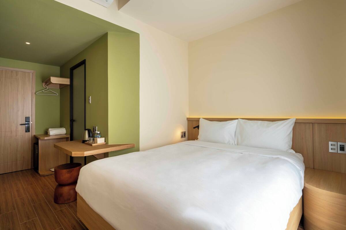 Une chambre d’hôtel avec un grand lit, un bureau en bois avec une chaise, un petit tabouret, une armoire en bois et un miroir au mur.