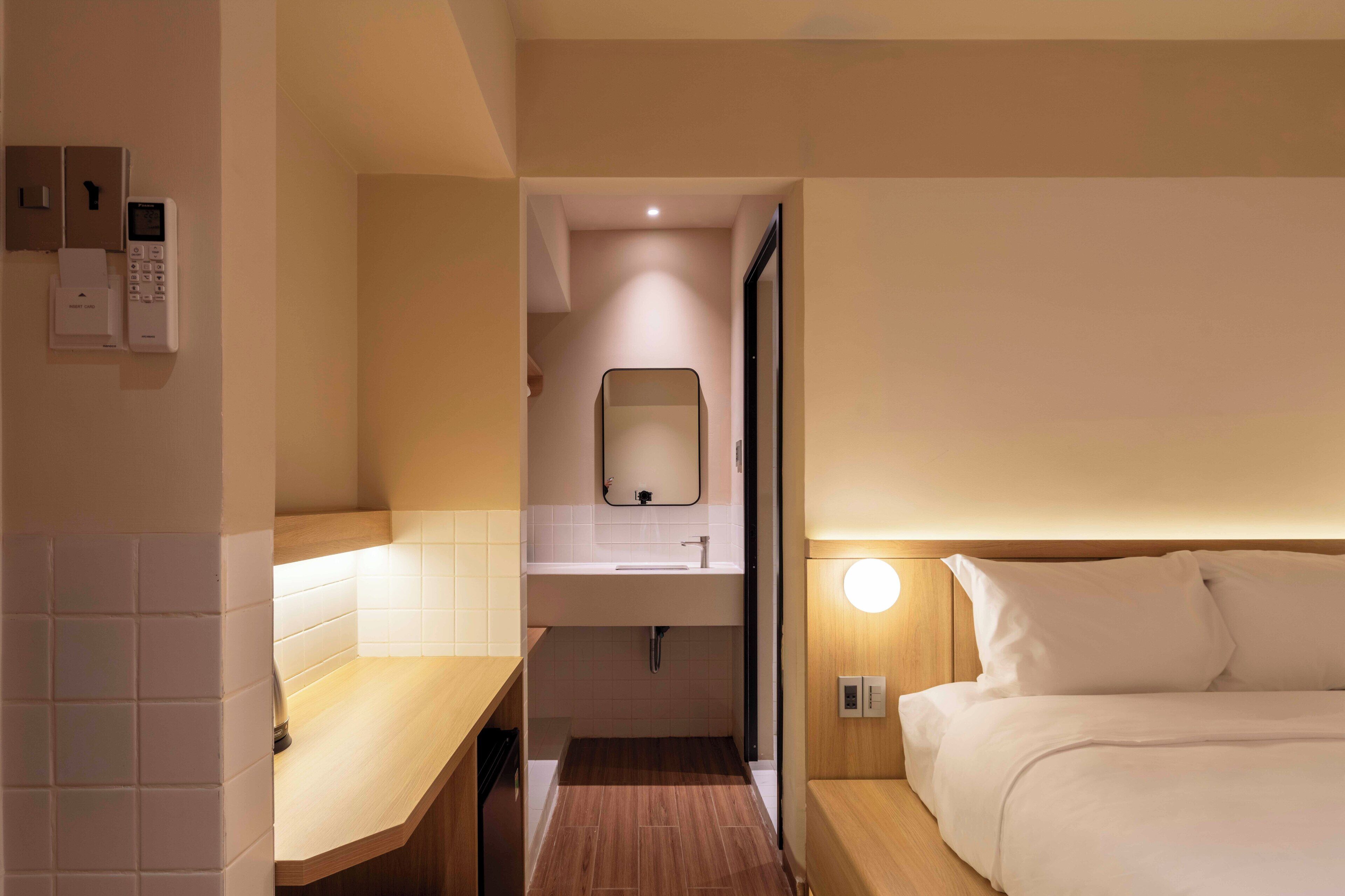 A hotel room with a wooden floor, a bed with white bedding, a bedside table, a small bathroom with a sink and mirror, and a wall-mounted phone.