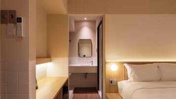Une chambre d’hôtel avec un plancher en bois, un lit avec une literie blanche, une table de chevet, une petite salle de bain avec un lavabo et un miroir, et un téléphone fixé au mur.