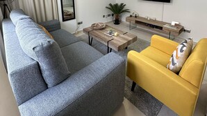 Een moderne woonkamer met een grijze modulaire bank, een gele fauteuil, een houten salontafel en een flatscreen televisie die aan de muur is bevestigd.