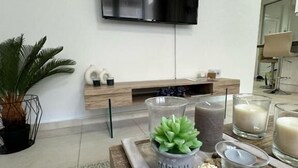 Een moderne woonkamer met een flatscreen televisie aan de muur, een houten salontafel met decoratieve objecten en een potplant.