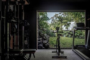 A gym with a treadmill, weight bench, and exercise equipment, with a view of a garden through a glass door. - Janjisurga by Ekosistem (Cepaka)