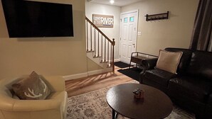 A living room with a sofa, a coffee table, a television mounted on the wall, a staircase, and a door.
