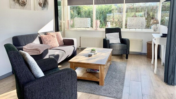 Una sala de estar moderna con un sofá gris, una mesa de centro de madera y un sillón negro. Hay dos ventanas con persianas, un radiador y una mesita con una lámpara.