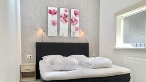 Un dormitorio moderno con cabecera negra, mesita de noche de madera, cama blanca con edredón y tres cuadros florales en la pared.