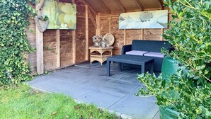 Un espacio de estar al aire libre con madera, que incluye una mesa y sillas, rodeado de follaje y un muro de madera decorado con obras de arte.