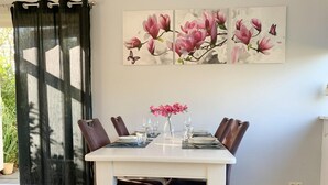 Un comedor con una mesa blanca dispuesta para cuatro personas, rodeada de sillas marrón oscuro. Un cuadro floral decora la pared sobre la mesa.