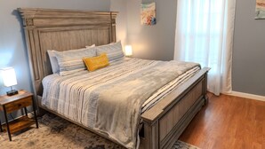 A bedroom with a wooden headboard, a bed with striped bedding, a small bedside table with a lamp, and a window with white curtains. - Harbor ViewCharming 3-bedroom house in delightful Acadia Area AC, WiFi (Ellsworth)