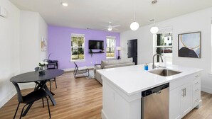 A modern kitchen with a white island, a dining area with a round table, and a living space with a sofa and a TV.
