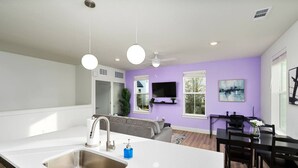 Una cocina moderna con una isla grande, una zona de comedor con mesa negra y una sala de estar con sofá gris y televisor.