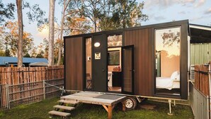 Eine kleine, moderne Hütte mit Bett, einer kompakten Küchenzeile und einer Terrasse mit Tisch und Stühlen.