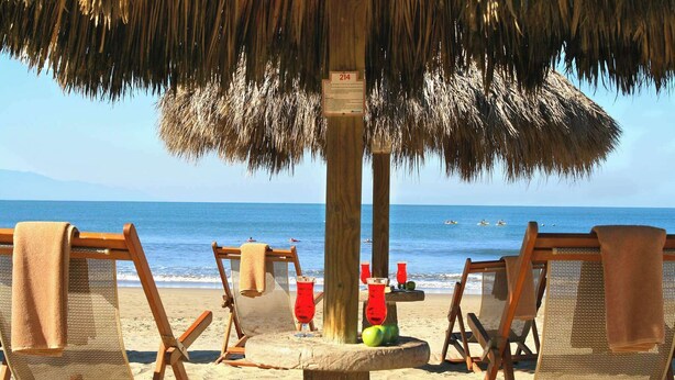 A beach with wooden chairs under a thatched umbrella, a sign with the number 214, and a view of the sea.