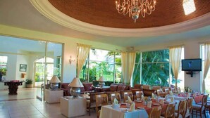 A spacious dining area with multiple tables and chairs, a chandelier, and large windows offering a view of greenery outside.