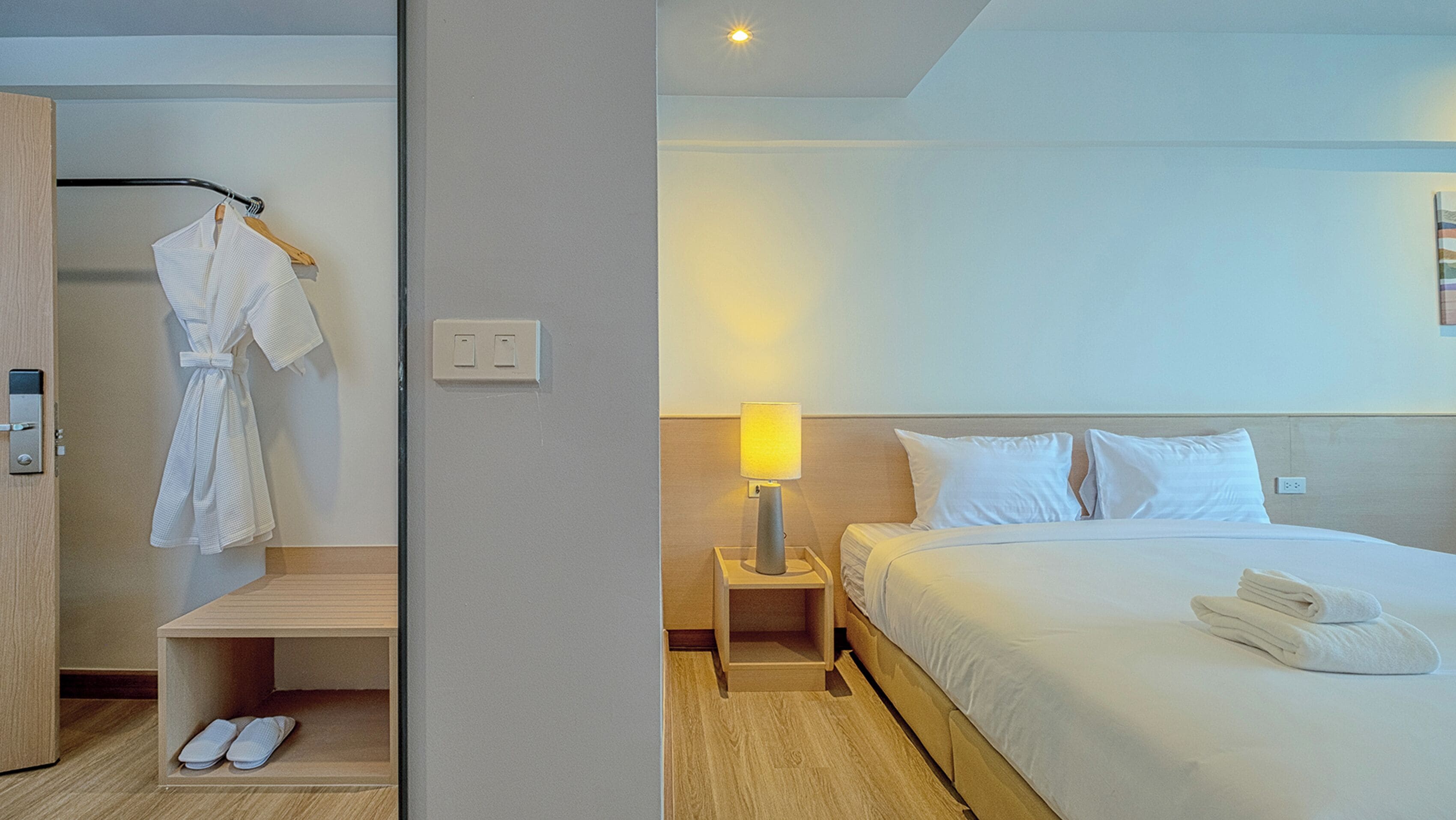 A hotel room with a bed, a nightstand with a lamp, a robe hanging on a rack, and a shoe rack.