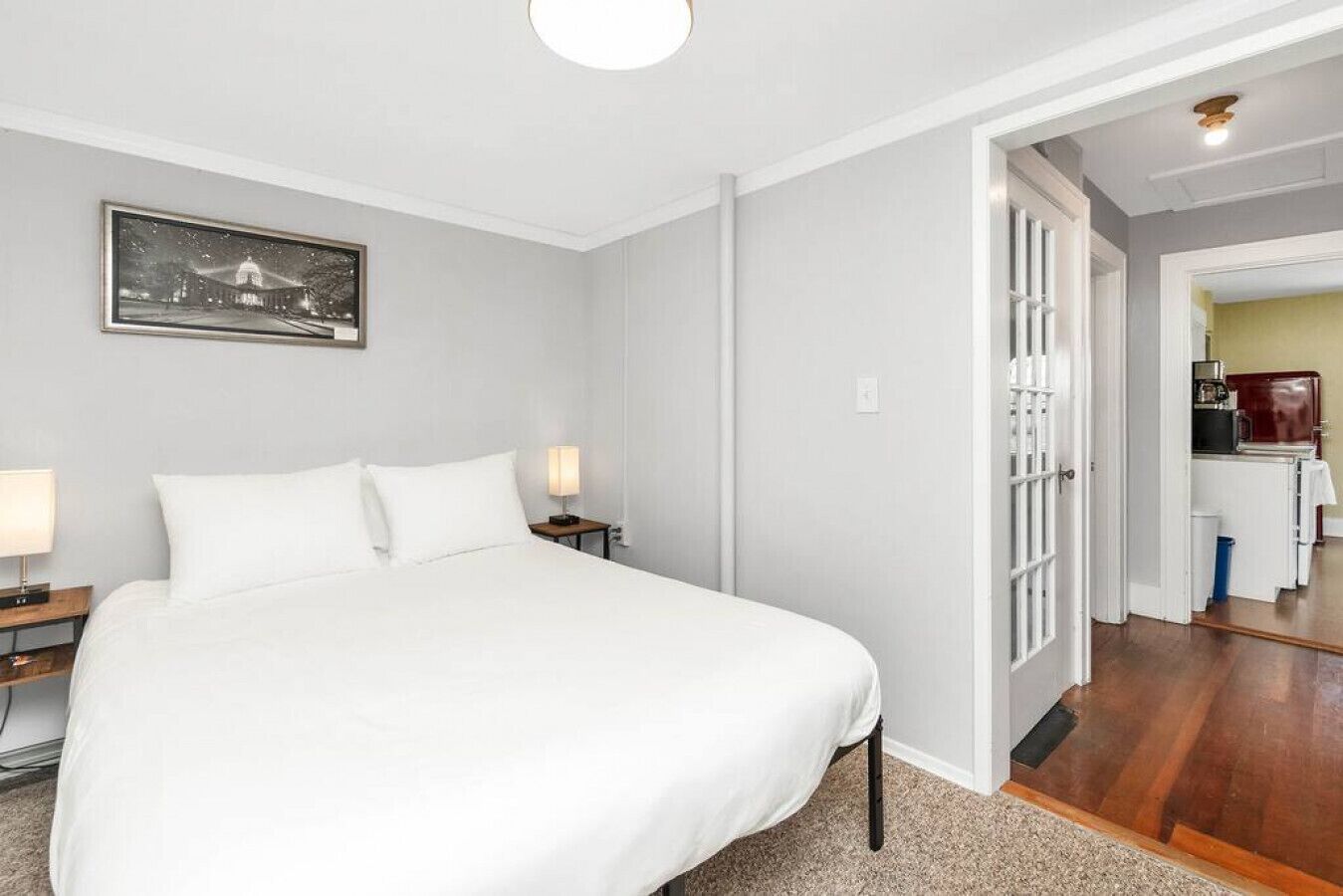 A hotel room with a large bed, bedside lamps, a framed picture on the wall, and a view into the kitchen area.