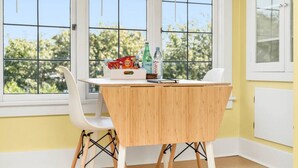 Un pequeño comedor con una mesa de madera y sillas blancas, una ventana con vistas a árboles, y una pared amarilla.