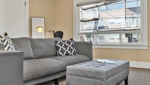 Una sala de estar con un sofá gris, almohadones estampados, una mesita auxiliar y una ventana con vista a un edificio.