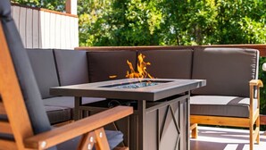 Una terraza de madera con fogata integrada, asientos acolchados y vistas a exuberante vegetación.