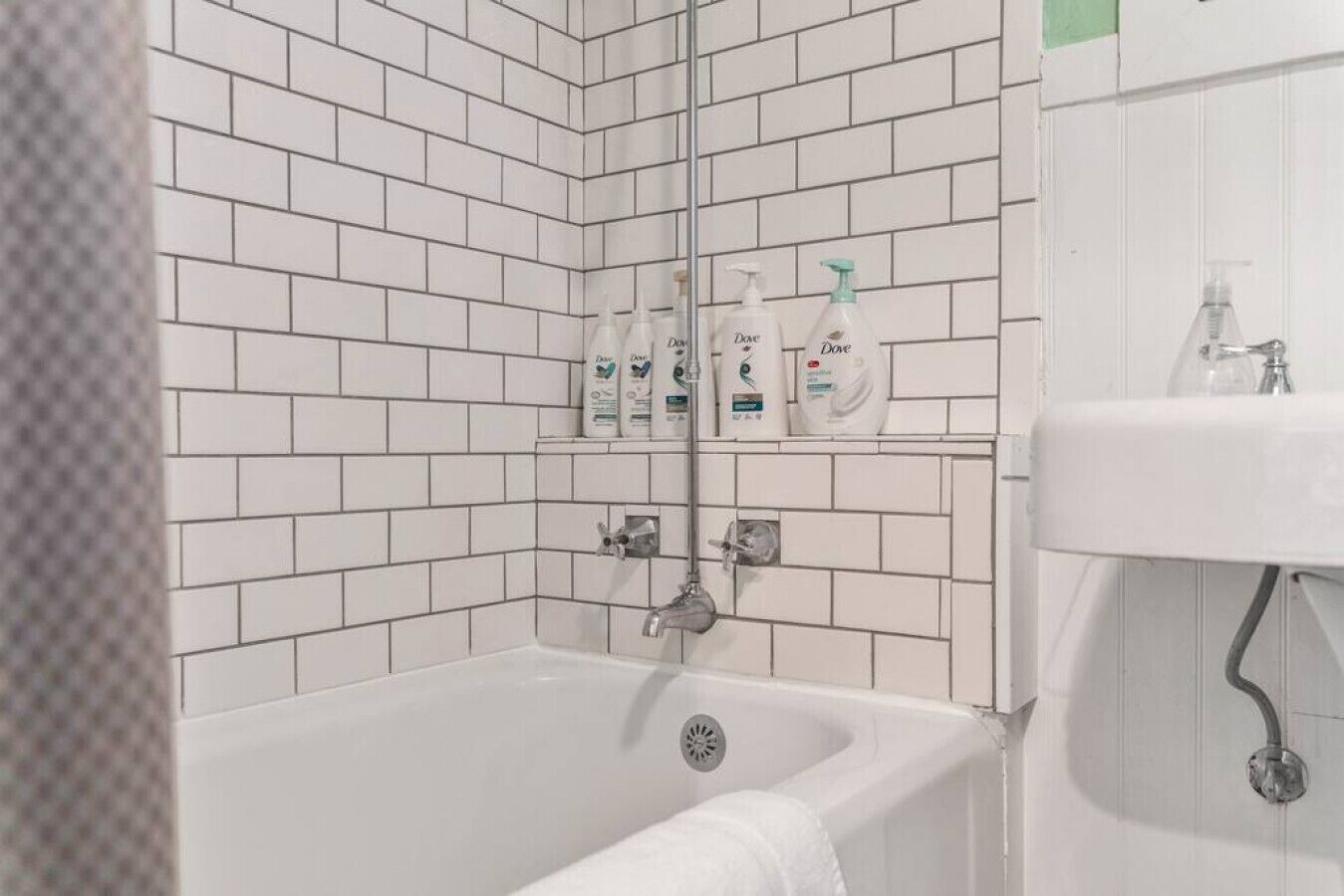 Une salle de bain avec des murs recouverts de carreaux blancs, une baignoire, un lavabo et trois flacons de savon pour les mains Dove sur une étagère.