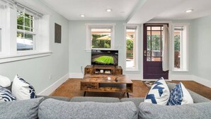 Ein Wohnzimmer mit Fernseher, Couchtisch aus Holz, grauem Sofa und einer Tür, die zu einem Außenbereich führt.