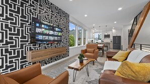 A modern living room with a black and white patterned wall, a flat-screen TV, leather sofas, a wooden coffee table, and a staircase. - Ballard Hangout - Arcade, Covered deck, Sleeps 10! (Seattle)