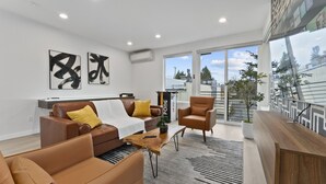 A modern living room with a brown leather sofa, a wooden coffee table, and a brown armchair. There are two abstract paintings on the wall, a potted plant, and a large window with a view of buildings. - Ballard Hangout - Arcade, Covered deck, Sleeps 10! (Seattle)
