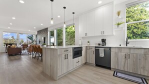 A modern kitchen with light wood cabinetry, a central island, and a dining area with a sofa and chairs. - Ballard Hangout - Arcade, Covered deck, Sleeps 10! (Seattle)