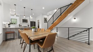 A modern kitchen with a wooden dining table, chairs, and a staircase with a metal railing. - Ballard Hangout - Arcade, Covered deck, Sleeps 10! (Seattle)