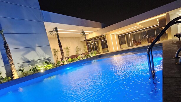 Una piscina con superficie de agua azul cristalina, rodeada por un edificio moderno con ventanas amplias y zonas de estar interiores.