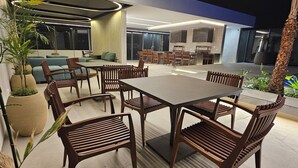 Un espacio moderno para comer al aire libre con una mesa redonda y sillas de madera, rodeado de plantas en macetas y una jardinera grande.