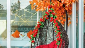 Una silla colgante de mimbre con un cojín rojo, rodeada de hojas otoñales y flores rojas, sobre césped artificial frente a un puerta de vidrio.