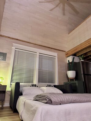 Ein Schlafzimmer mit Bett, Fenster mit Jalousien, einem Topf mit Pflanze und einem Kühlschrank.