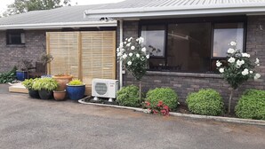 A residential building with a brick exterior, a wooden sliding door, and a window with white flowers. There are potted plants and a small garden area in front.