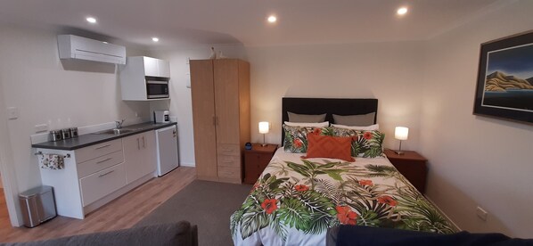 Un dormitorio compacto con cama, mesitas de noche, armario, kitchenette y aire acondicionado instalado en la pared.