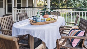 A wooden deck with a table set for two, featuring a white tablecloth, wicker basket with lemons, bottles, and a potted plant.