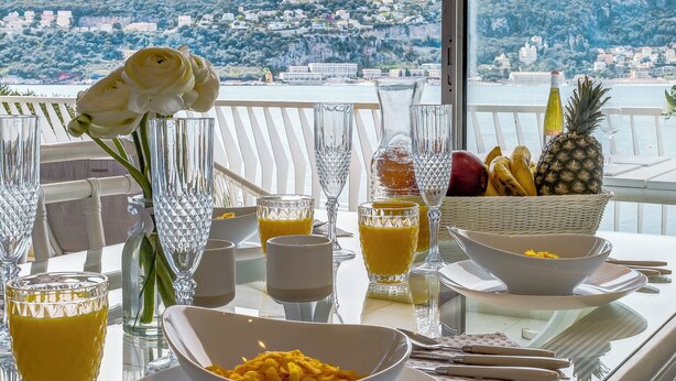 A table set with breakfast items, including fruits, a flower, and glasses, overlooking a scenic view.