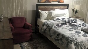 A bedroom with a bed, a red chair, a wooden nightstand, a lamp, and a painting of a beach scene. - Charming 2-bedroom apartment in Pleasant Valley Stream with AC, WiFi (Valley Stream)