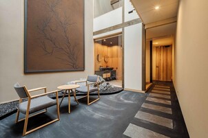 Un interior moderno con una zona de estar que incluye dos sillas y una mesita, un cuadro abstracto de gran tamaño en la pared, y una escalera que sube a otra habitación.