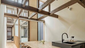 Une cuisine moderne avec des poutres apparentes, une longue table en bois entourée de chaises, un îlot central noir doté d’un évier et une vue dégagée grâce à de grandes fenêtres.