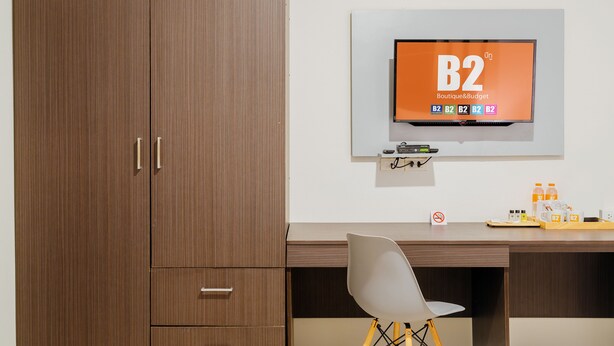 A modern room with a wooden wardrobe, a desk with a chair, a mounted TV displaying 'B2 Boutique&Budget', and a shelf with bottles and a tray. - B2 Chiang Rai Resort Boutique & Budget Hotel (Chiang Rai)