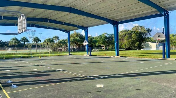 An outdoor basketball court with a covered seating area, surrounded by trees and a clear sky.