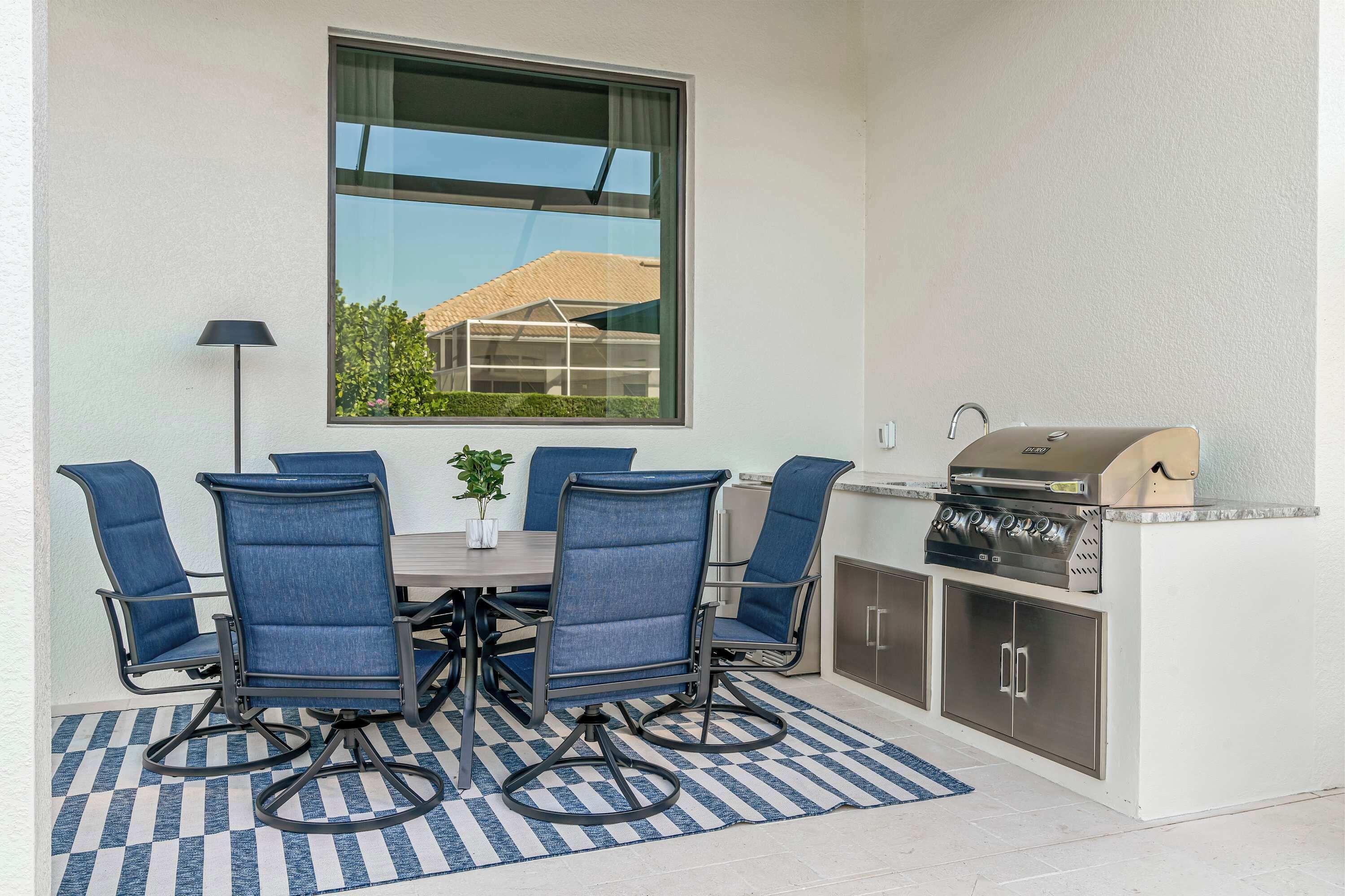 A patio with a table and chairs, a grill, and a view of a building through a window.