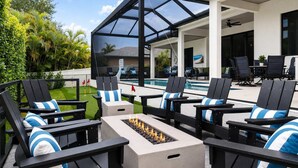 An outdoor patio with black chairs and blue-striped cushions, a fire pit, and a pool in the background. - Luxury 5BR - Pool, Game Room, Mini Golf, Fire Pit (Marco Island)