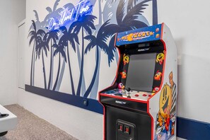 An arcade machine with a Pac-Man theme, located in a room with a wall mural of palm trees and a neon sign. - Luxury 5BR - Pool, Game Room, Mini Golf, Fire Pit (Marco Island)