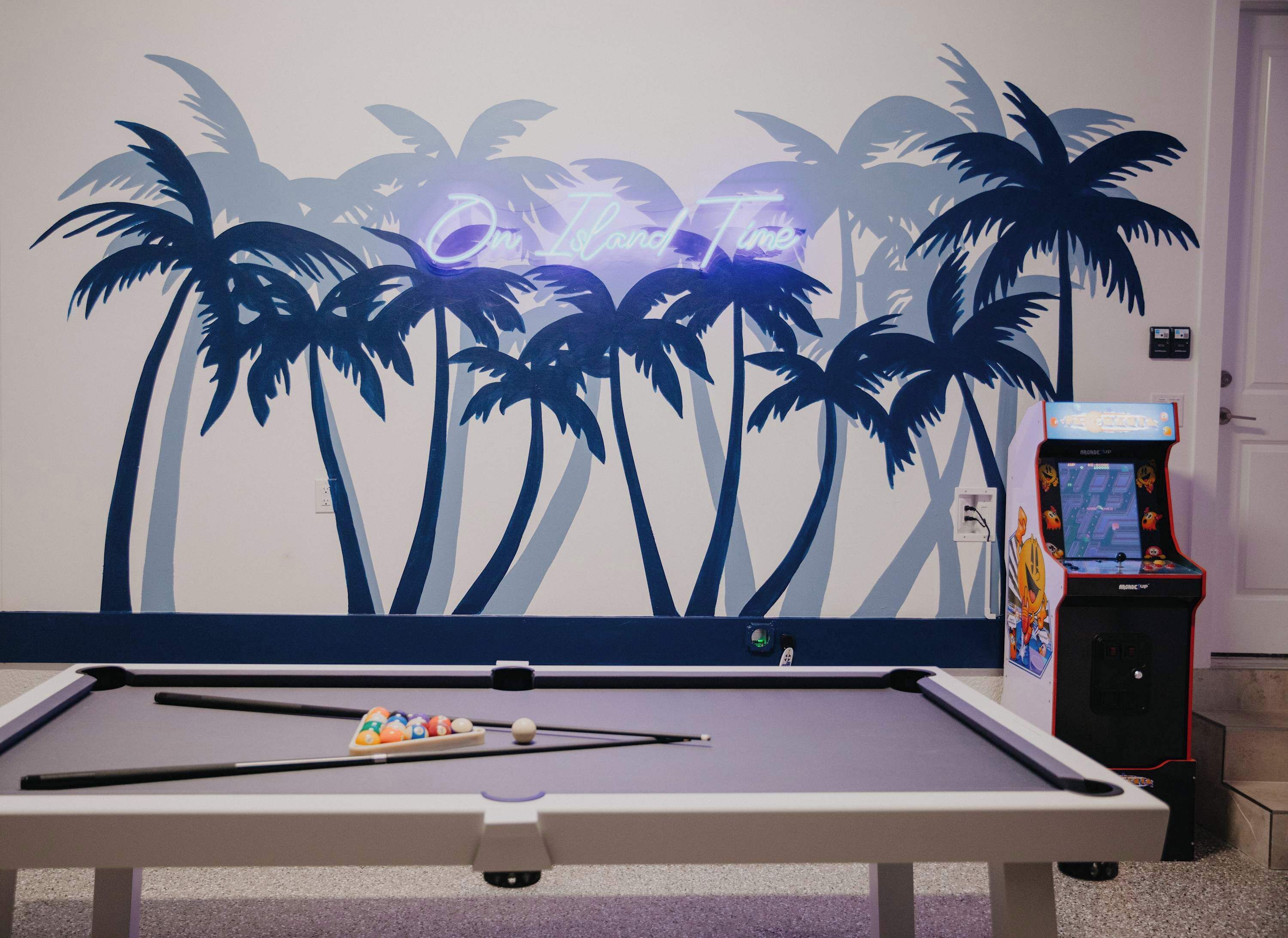 A pool table with a neon sign reading "One Island Time" and a pool table in a room with a wall mural of palm trees.